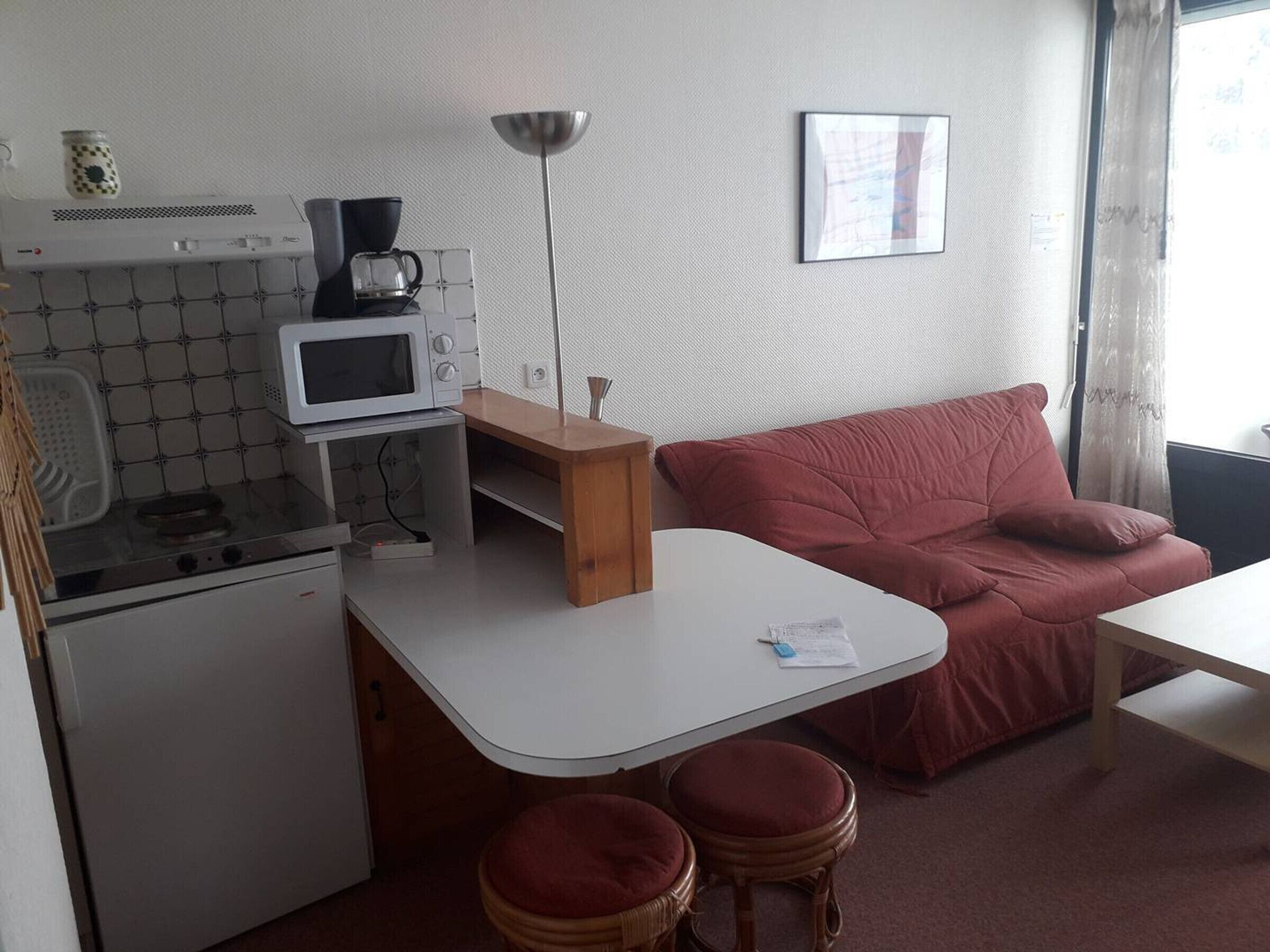 Een compact keukenhoekje met een magnetron, fornuis en koffiezetapparaat. Er staat een klein tafeltje met krukken en een rode bank. Op het tafeltje staat een lamp, en aan de muur hangt een lijst met een foto.
