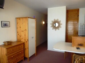 A small, compact room with a wooden wardrobe, a small table with a chair, and a mirror on the wall.