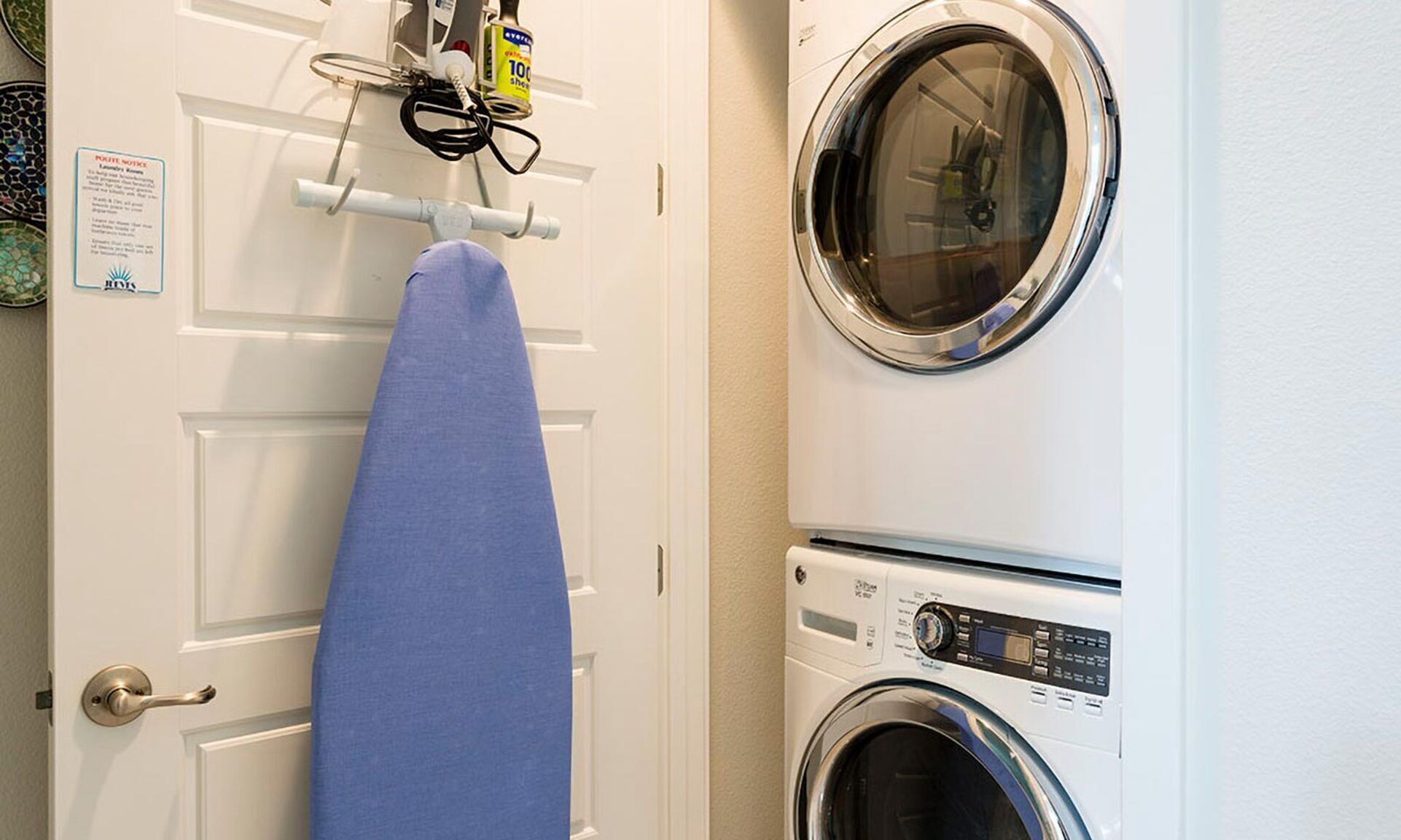 A laundry area with a washing machine and dryer, a blue ironing board, and a notice on the door.