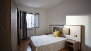A bedroom with a bed, a nightstand, a bedside table, a window with curtains, and a view of buildings outside.