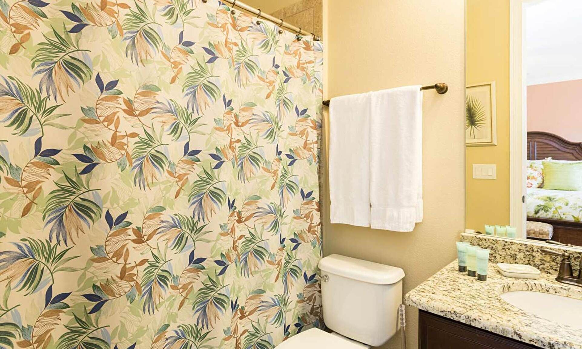 A bathroom with a shower curtain featuring a tropical leaf pattern, a white toilet, a sink with a granite countertop, and a mirror reflecting a bed with floral bedding.