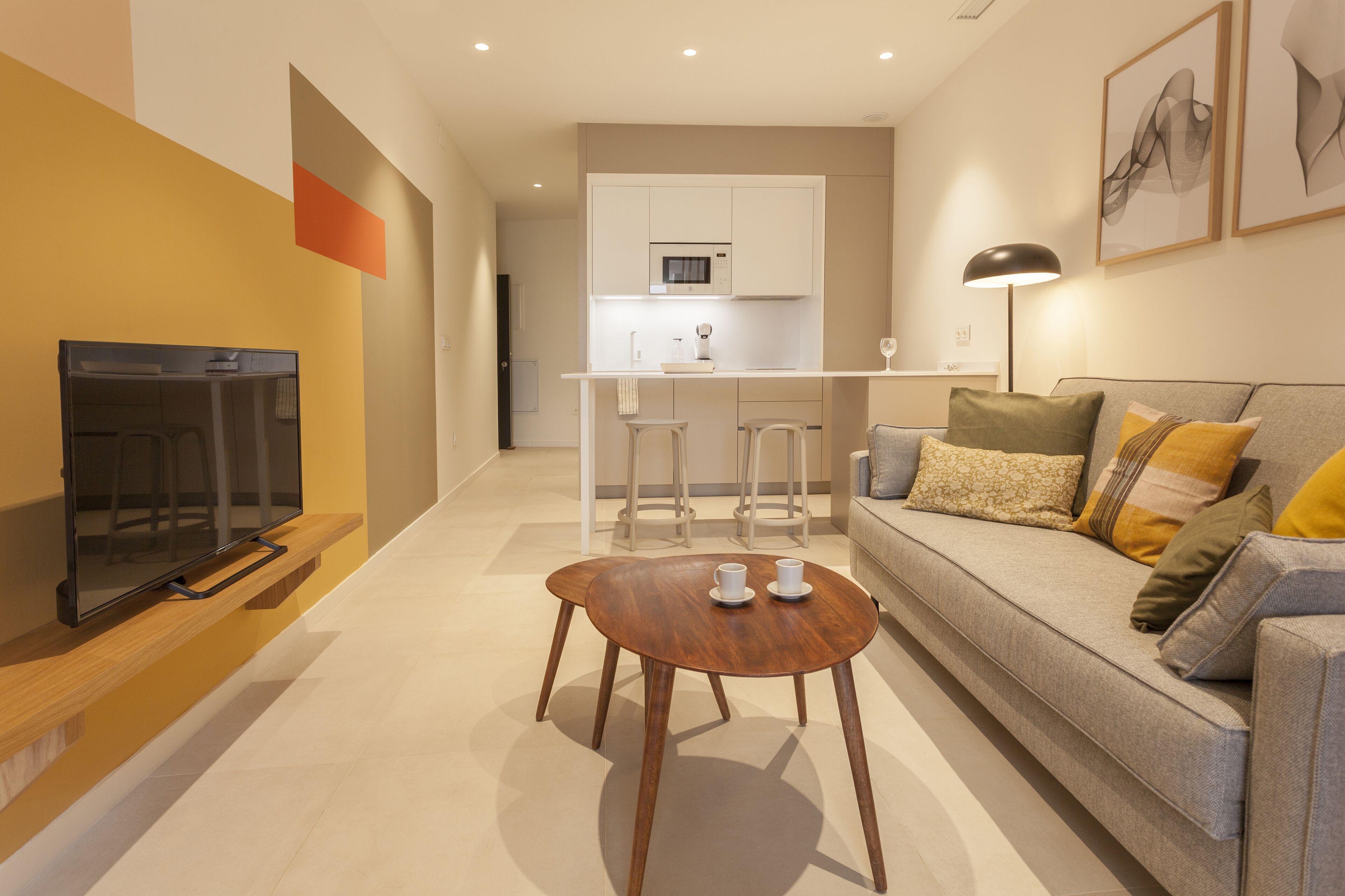A modern living room with a flat-screen TV, a wooden coffee table, and a sofa with cushions. There is a kitchen area in the background with white cabinets and a microwave.