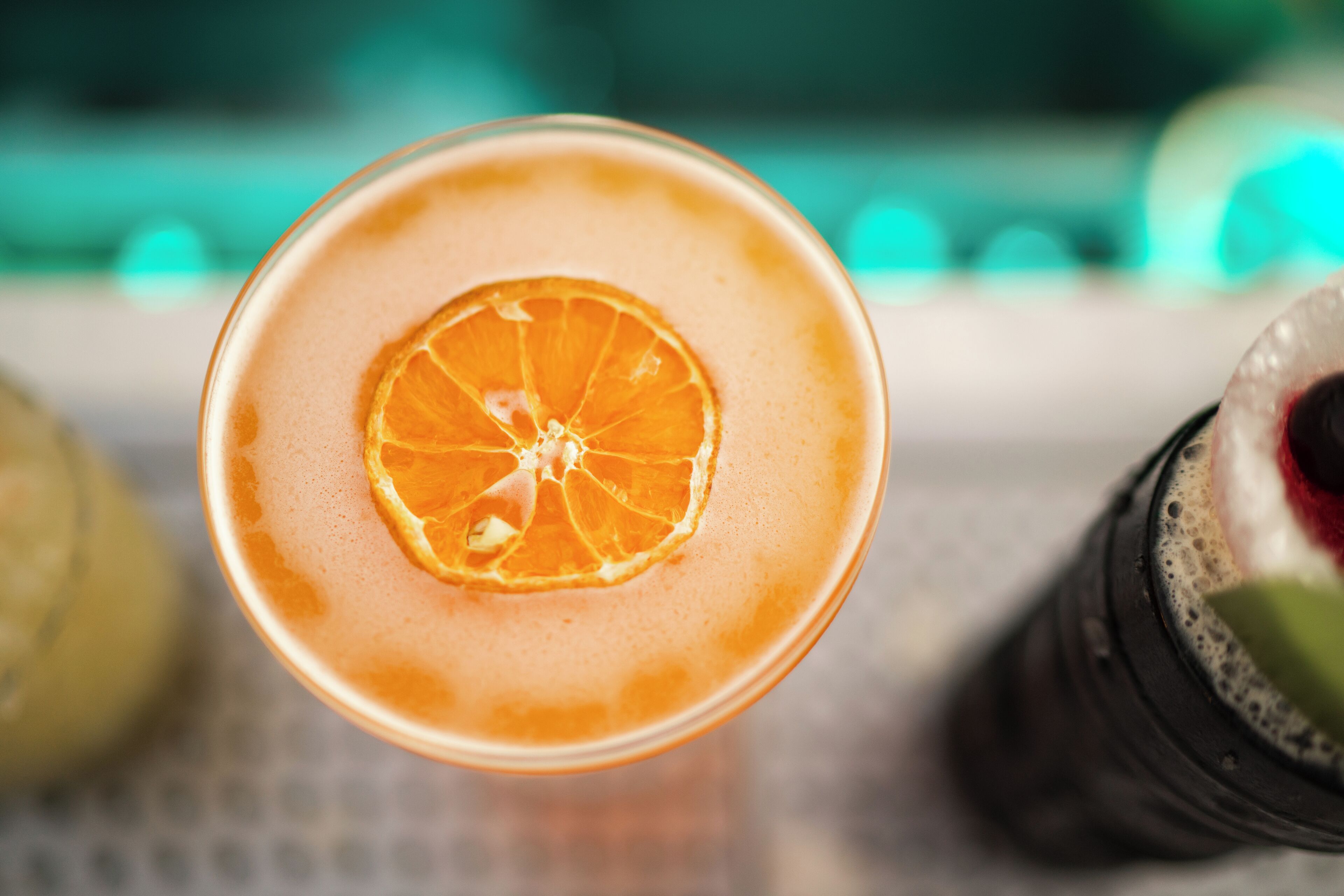 Un vaso de bebida anaranjada con una rodaja de naranja en el borde, sobre una superficie con fondo desenfocado.