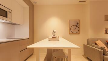 A modern kitchen with a white countertop, a built-in microwave, and a coffee machine. There is a white dining table with two stools and a grey sofa with cushions. A framed bicycle artwork is on the wall.