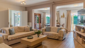 Un salon spacieux, doté d’un canapé beige, d’une table basse en bois et offrant une vue sur l’extérieur grâce à de grandes fenêtres.