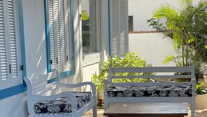 A white bench with a black and white patterned cushion, positioned on a brick-paved patio adjacent to a building with blue trim and shutters. - Casa na Praia do Canto - 6 Quartos - Búzios (Armação dos Búzios)