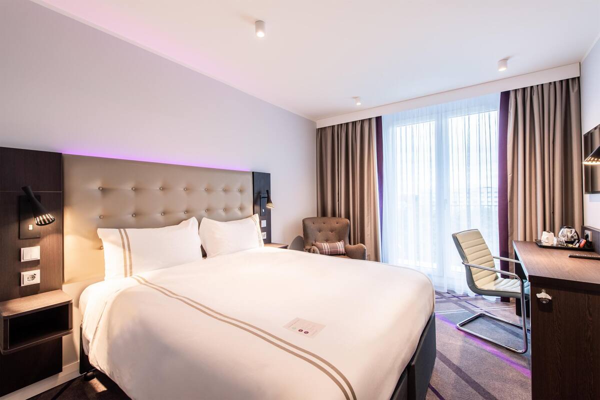 A hotel room with a large bed, a desk, a chair, a nightstand, and a wall-mounted lamp.