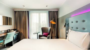 A hotel room with a bed, a desk, a chair, a lamp, and curtains.