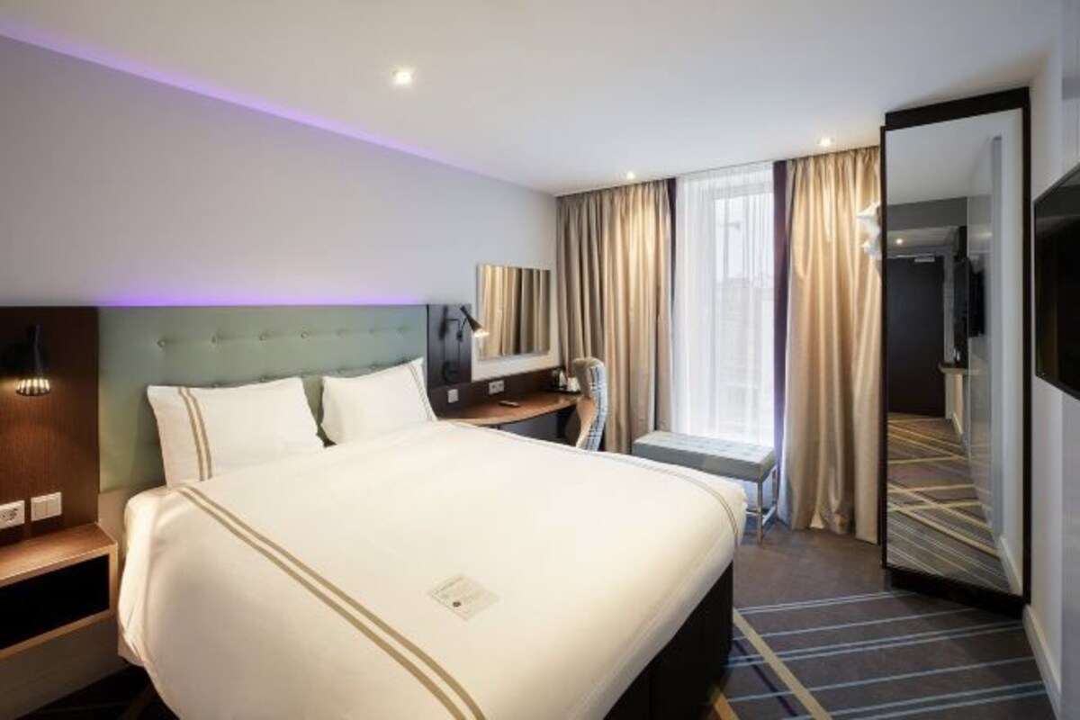 A hotel room with a large bed, a bedside table, a mirror, a TV, and a window with curtains.
