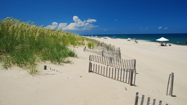 A beach with a wooden fence, beach umbrellas, and a clear blue sky. - Mariner's Star Five Bedroom Home in Nags Head with Private Pool and Hot Tub (Nags Head)