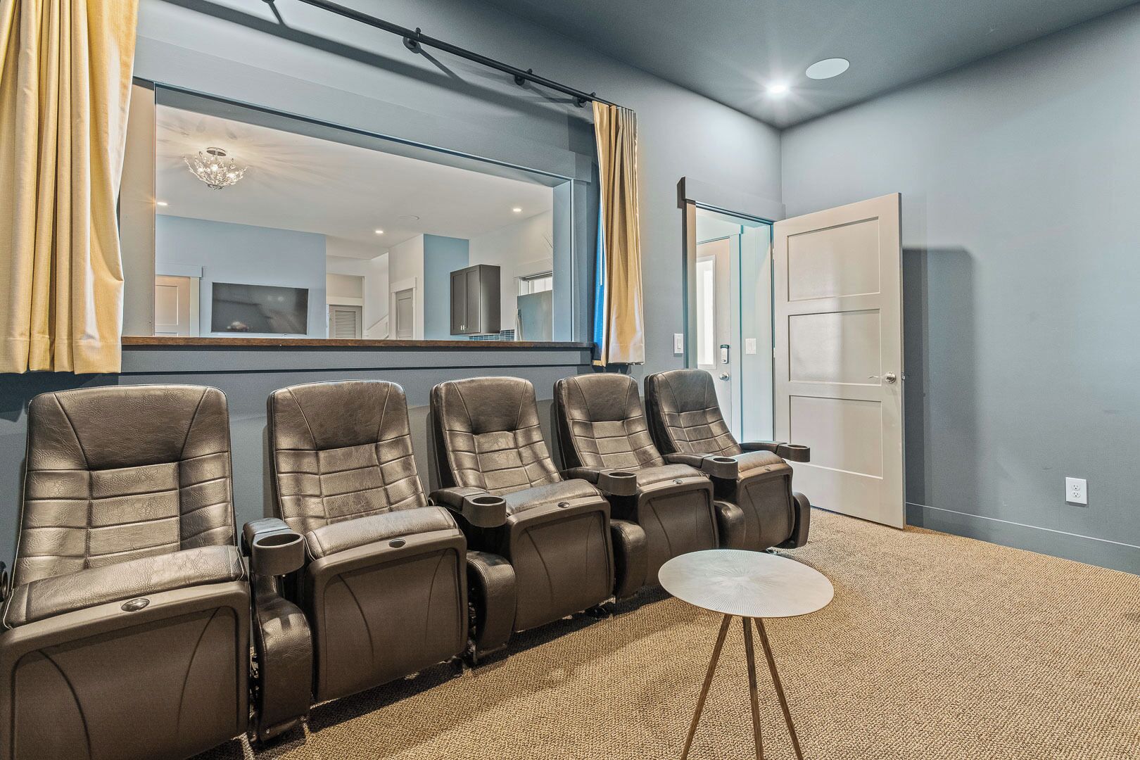 A room with four massage chairs, a small round table, and a mirror reflecting a kitchen area.