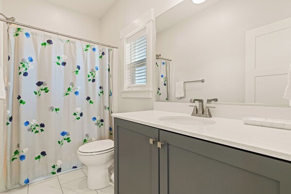 A bathroom with a shower curtain featuring a floral pattern, a toilet, a sink with a mirror, and a window with blinds.