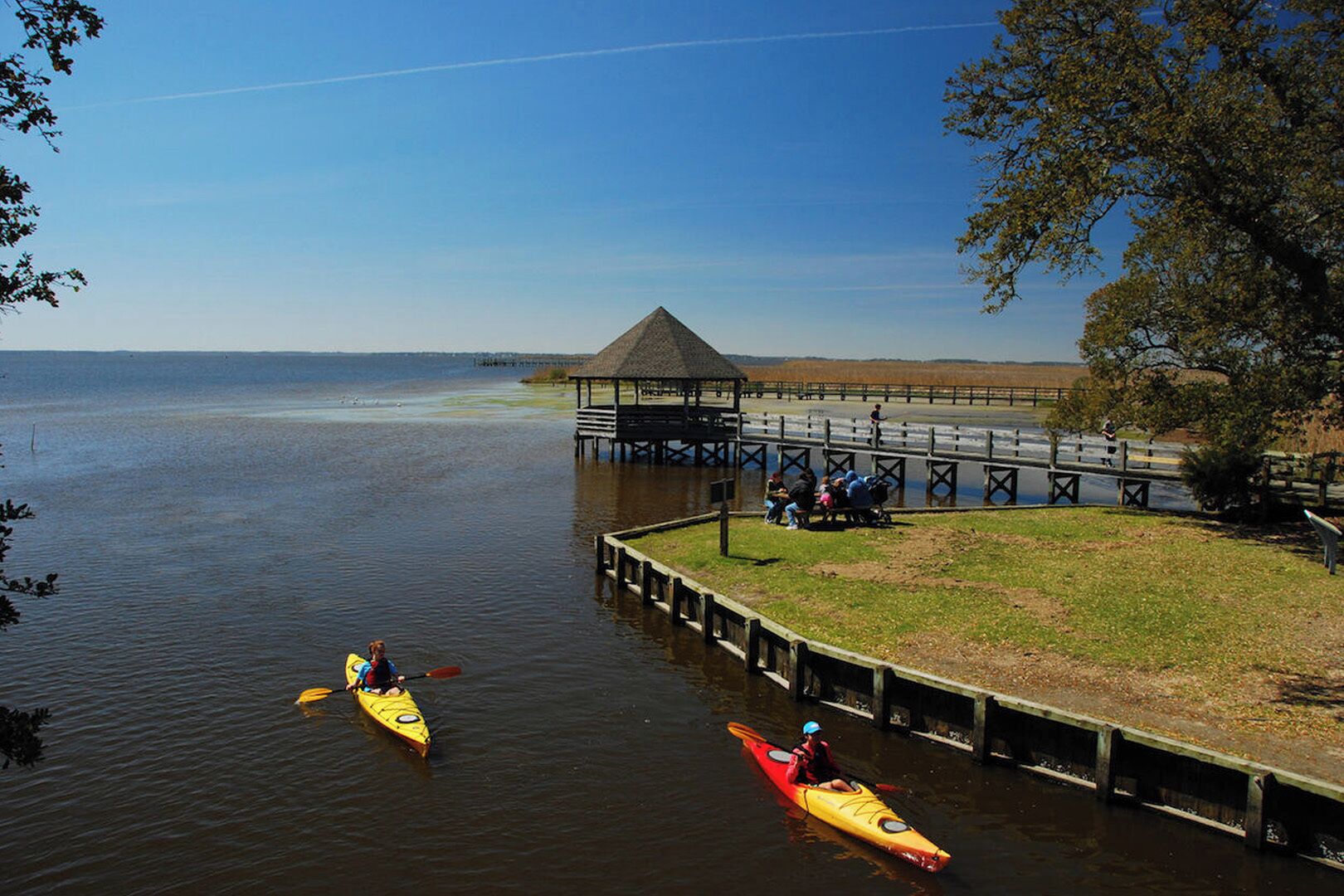 A lakeside pier with a gazebo, kayaks, and people sitting on benches.