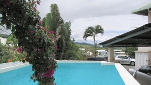 A swimming pool with a stone border, surrounded by greenery and a covered area with parked cars. - F3 apartment in the heart of Petite Ile (Saint-Pierre)