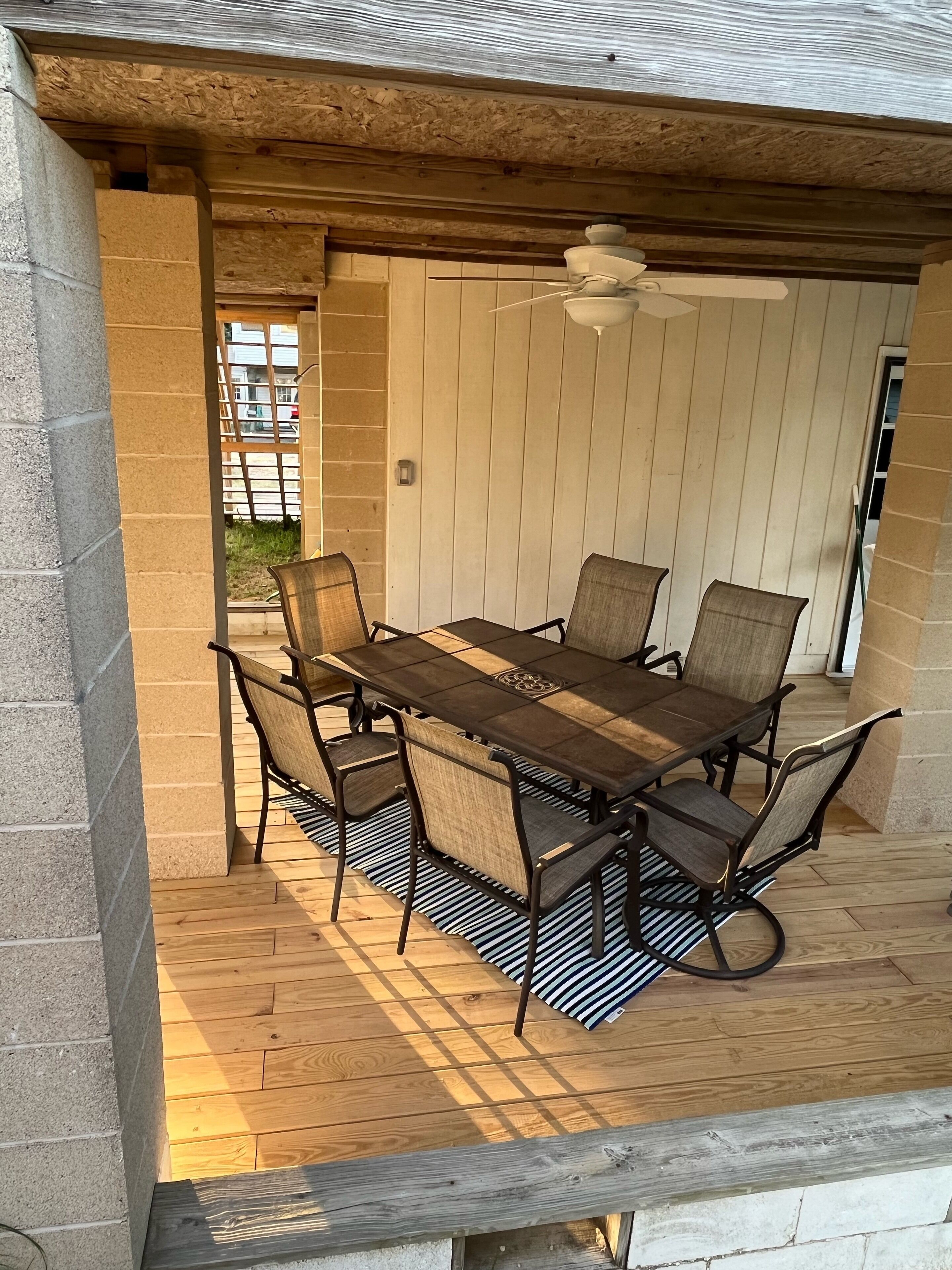 Een overdekt terras met een houten tafel en stoelen, een plafondventilator en uitzicht op de omgeving.