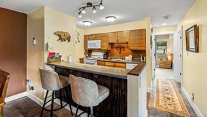 Una cocina con una isla central, armarios de madera y un microondas. Hay dos taburetes de bar, una alfombra y una decoración de oso en la pared.