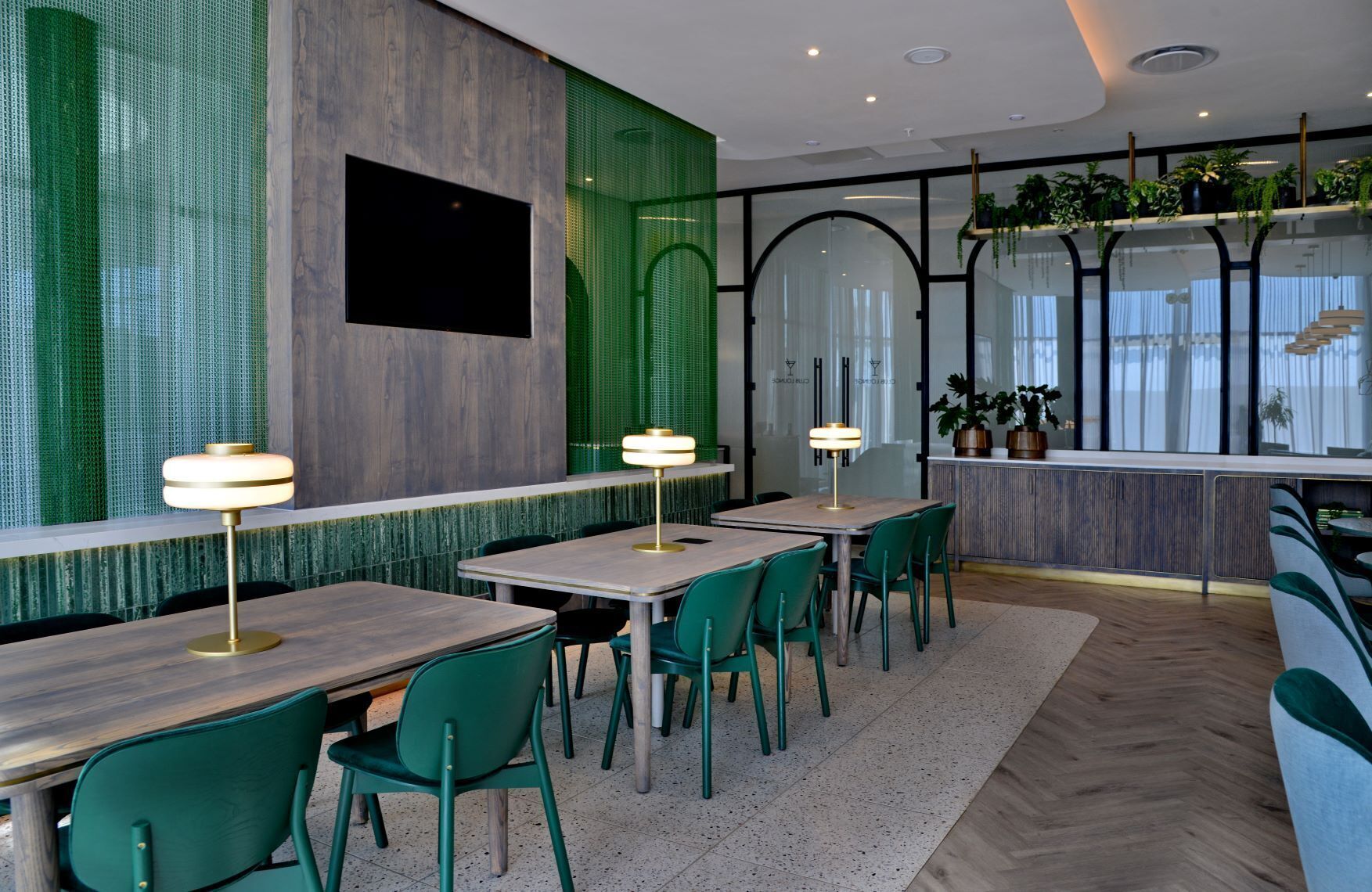 Een modern restaurantinterieur met groene stoelen, houten tafels en een groen wandpaneel.