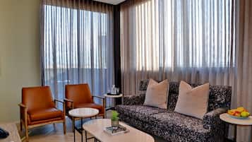 A modern hotel room with a sofa, two armchairs, a coffee table, and a small side table with a fruit bowl.