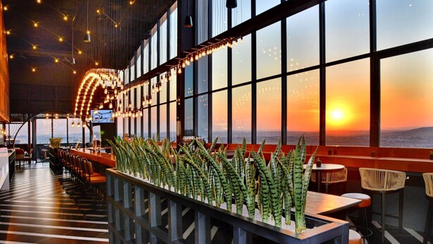 Un restaurante con una barra larga, sillas y mesas. Se ven grandes ventanales con vistas al atardecer.
