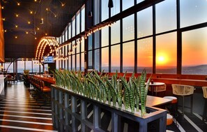 Un restaurante con una barra larga, sillas y mesas. Se ven grandes ventanales con vistas al atardecer.