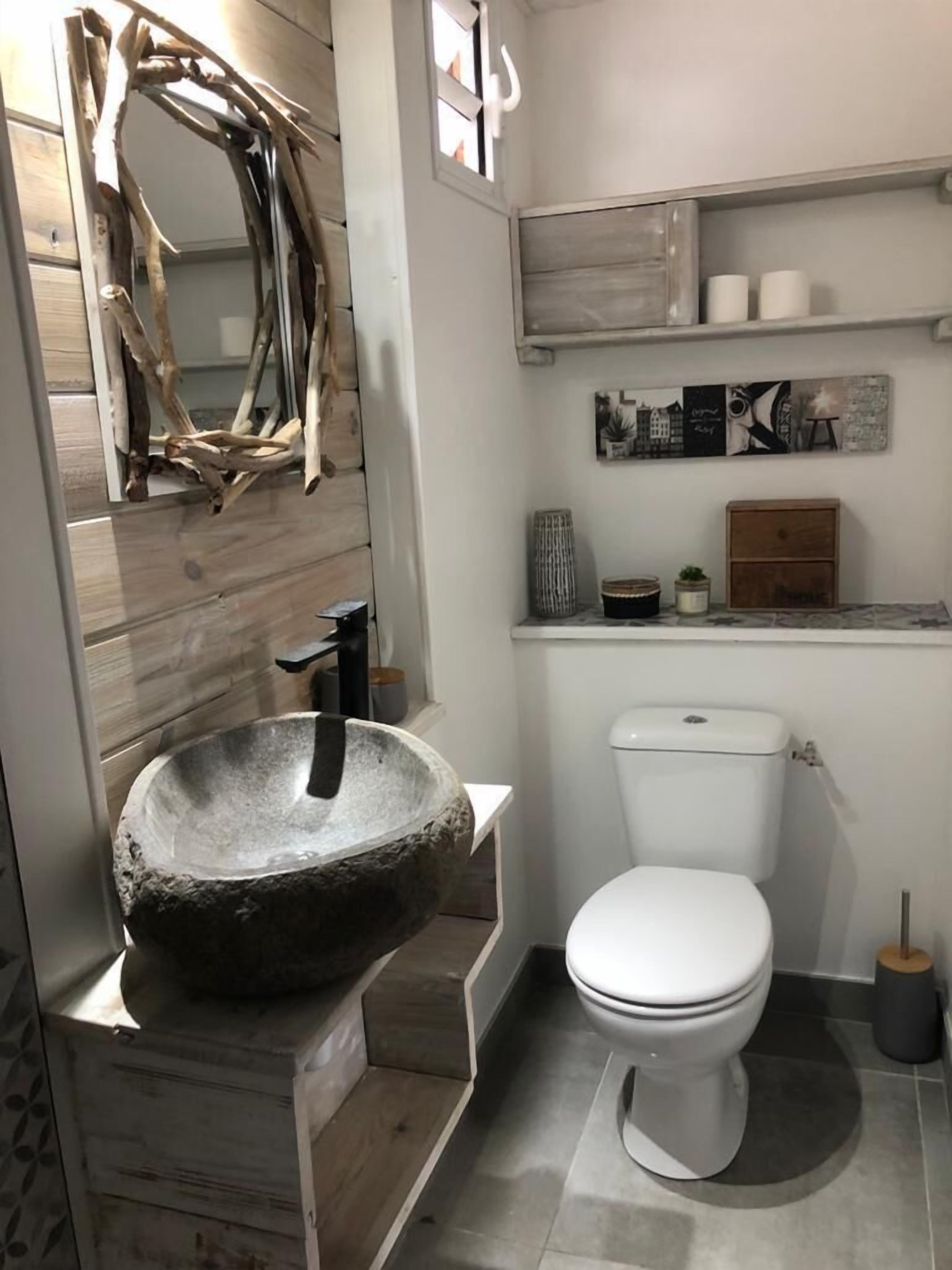 Une salle de bain avec un lavabo en pierre, des toilettes blanches, un lambris mural en bois et un miroir doté d’un cadre rustique.