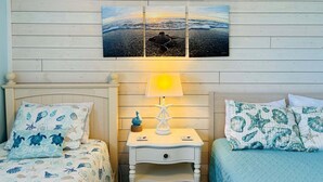 Quarto com duas camas, uma mesa de cabeceira com abajur e um quadro na parede retratando uma praia.