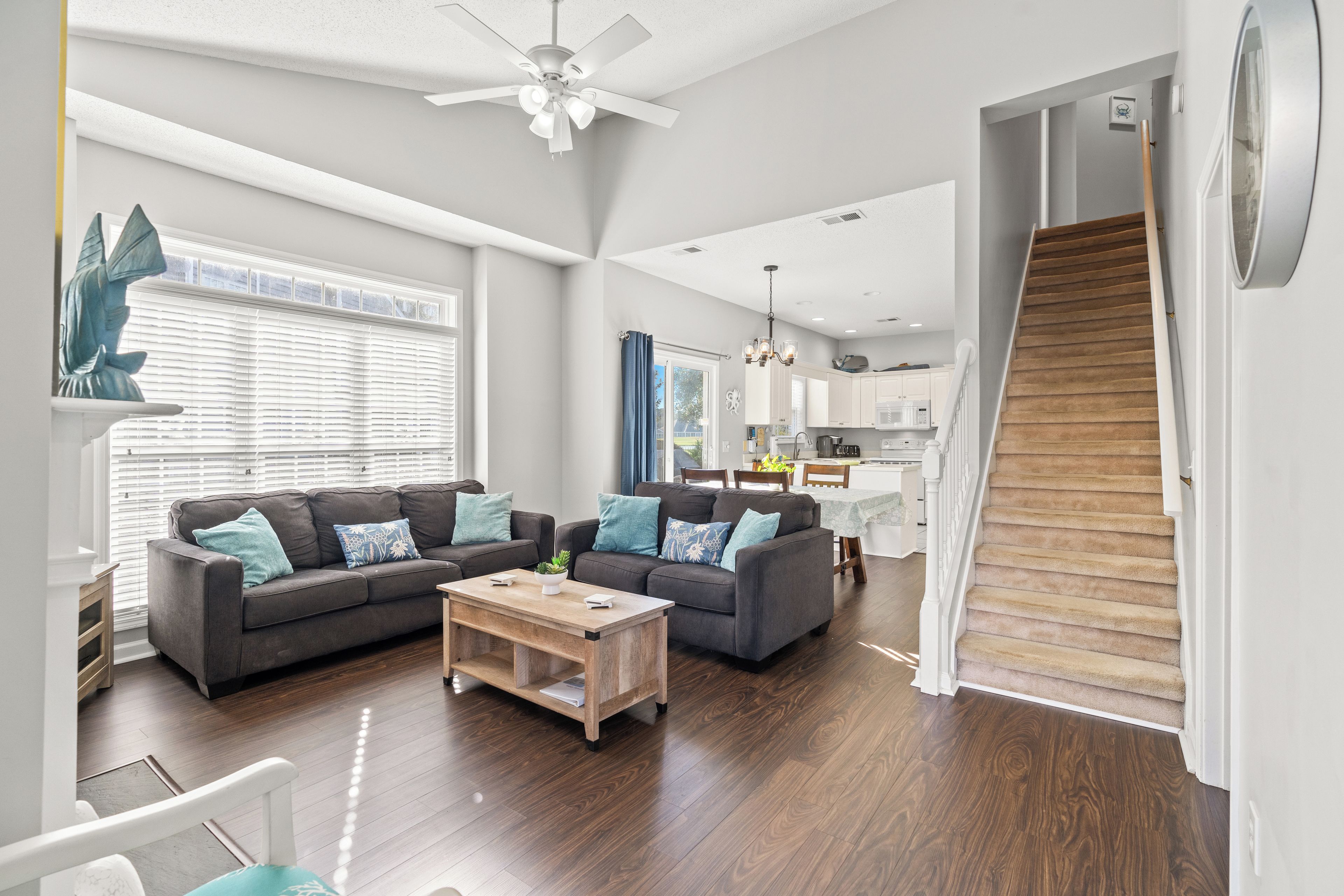 A spacious living room with a dark wood floor, two grey sofas with blue cushions, a wooden coffee table, and a ceiling fan. A staircase with wooden steps leads to an upper level.