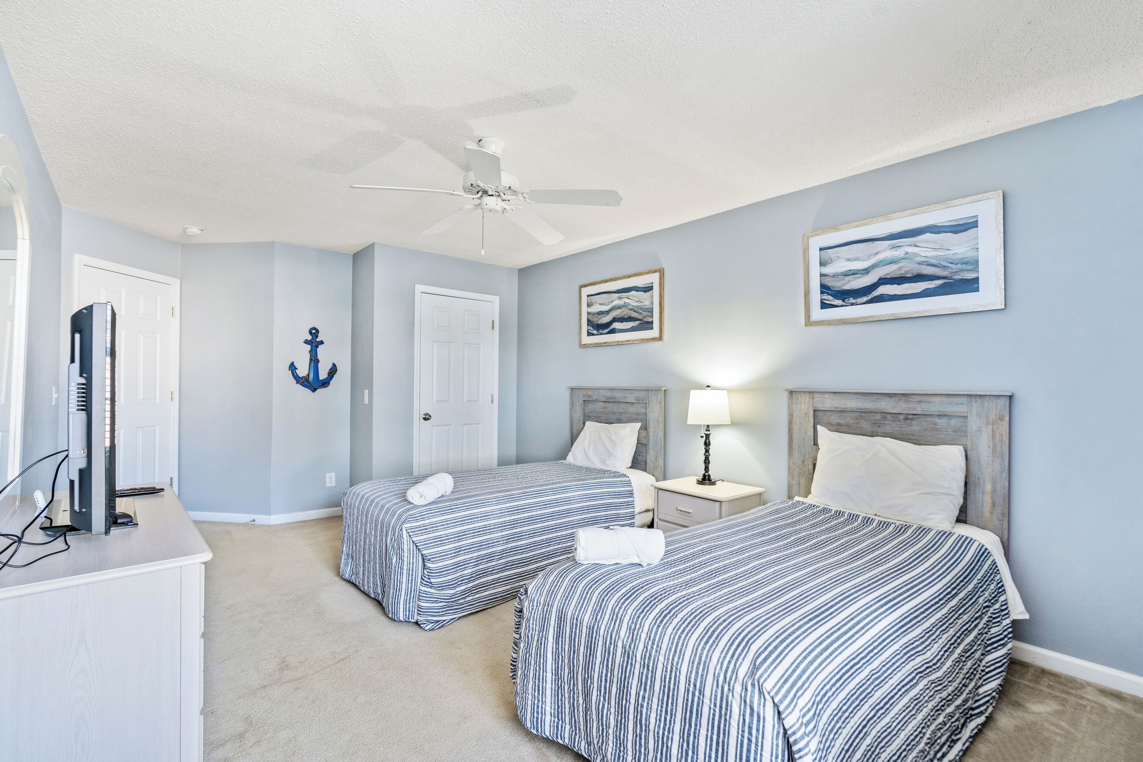A hotel room with two beds, a desk, a ceiling fan, and artwork on the walls.