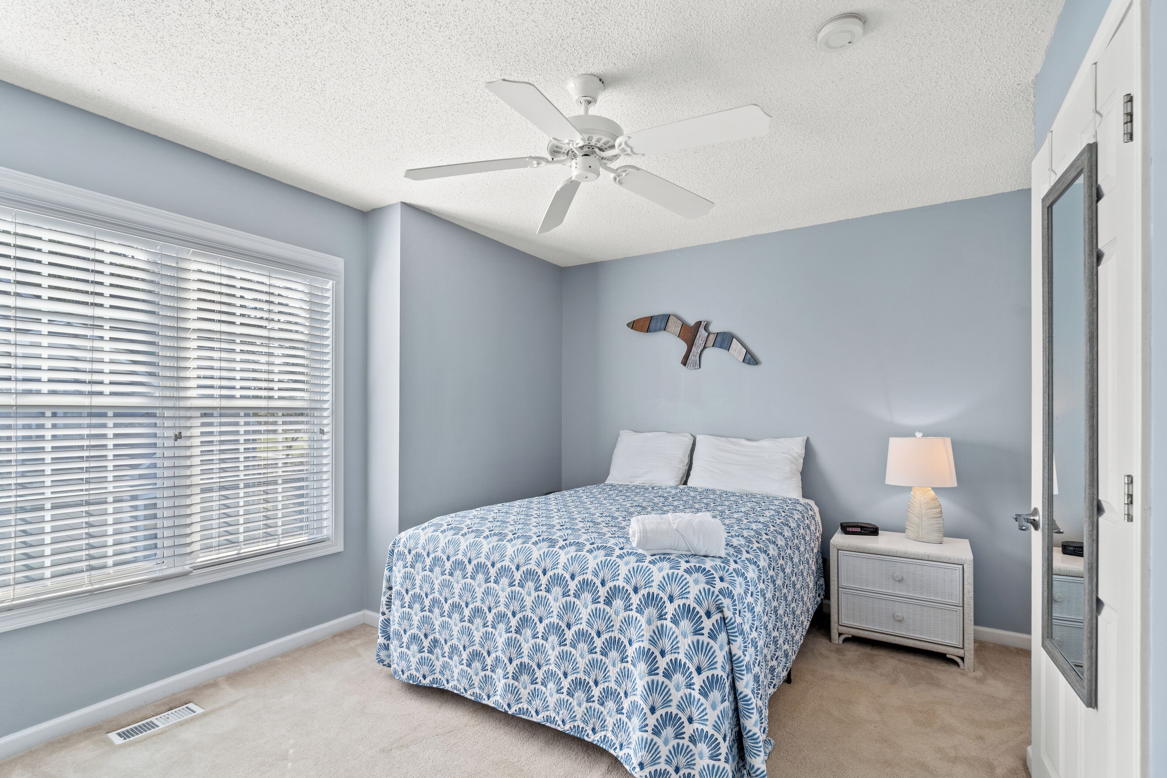 A bedroom with a bed, a nightstand, a ceiling fan, a window with blinds, and a wall-mounted bird decoration.