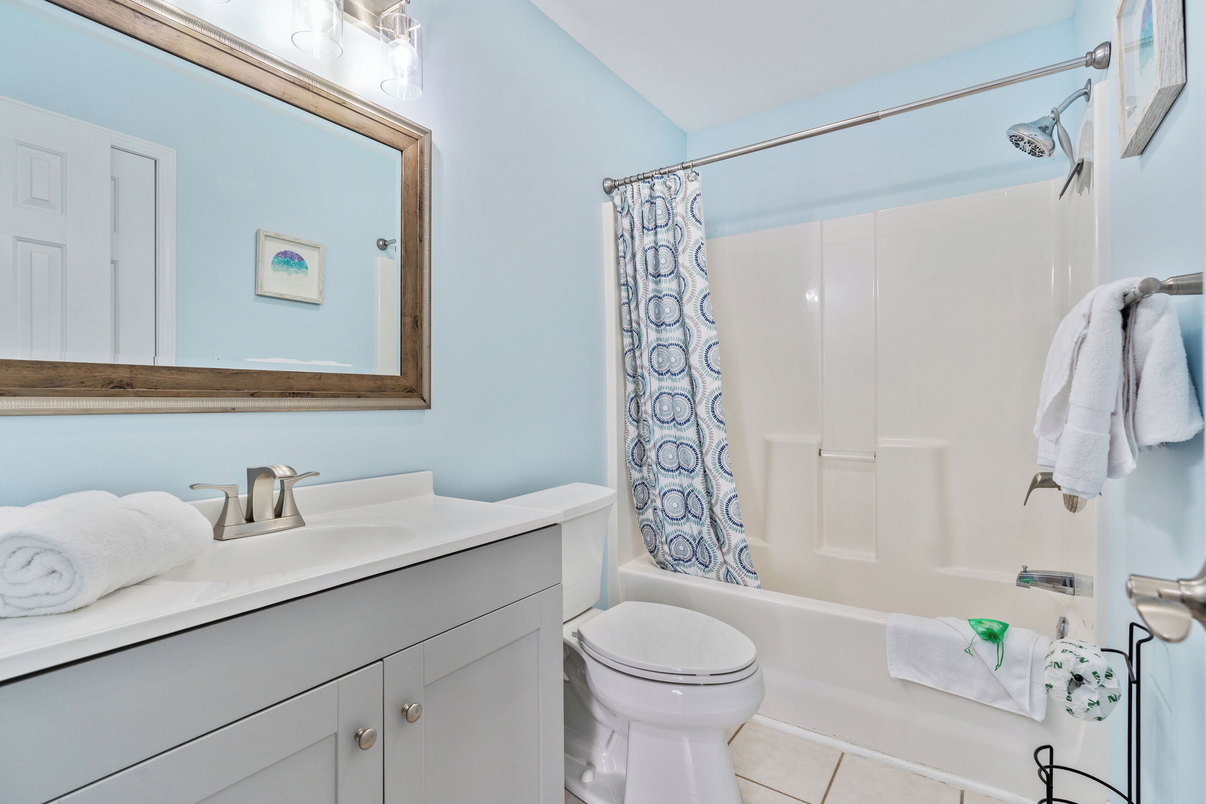Un baño con lavamanos, inodoro y bañera blancos. Se observa una cortina de ducha con estampado azul y blanco. Hay un perchero con toallas.