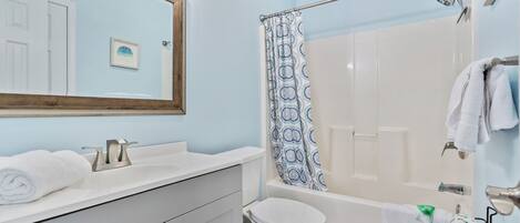 Un baño con lavamanos, inodoro y bañera blancos. Se observa una cortina de ducha con estampado azul y blanco. Hay un perchero con toallas.