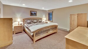 Quarto com cama de madeira, duas mesas de cabeceira com abajures, um guarda-roupa, uma janela com vista e iluminação embutida no teto.