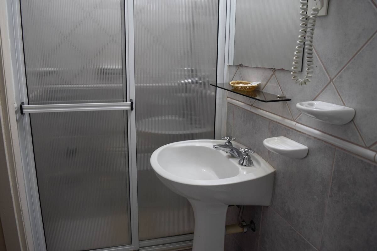 Un baño con cabina de ducha de vidrio, un lavabo blanco con plato para jabón y un teléfono fijo en la pared.