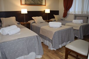 Habitación de hotel con tres camas individuales, cada una con una almohada y una toalla enrollada. Hay dos mesitas de noche con lámparas, una silla de madera y una ventana con cortinas.