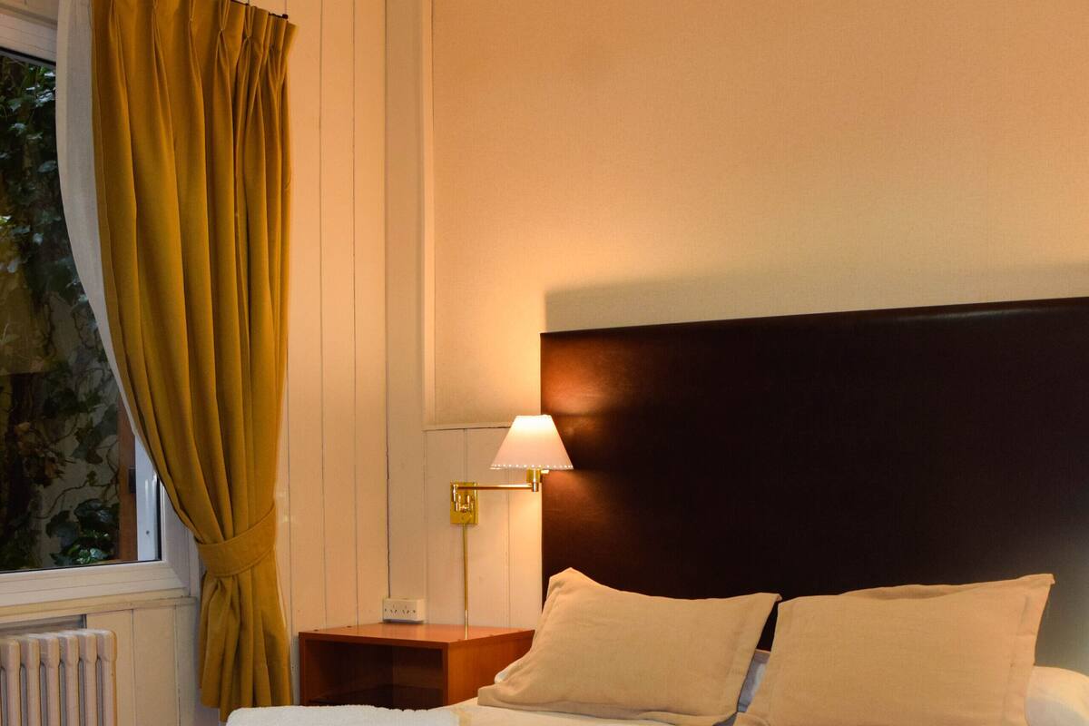 Una cama bien hecha con sábanas blancas y dos toallas enrolladas, una mesita de noche con lámpara, y una ventana con cortinas.
