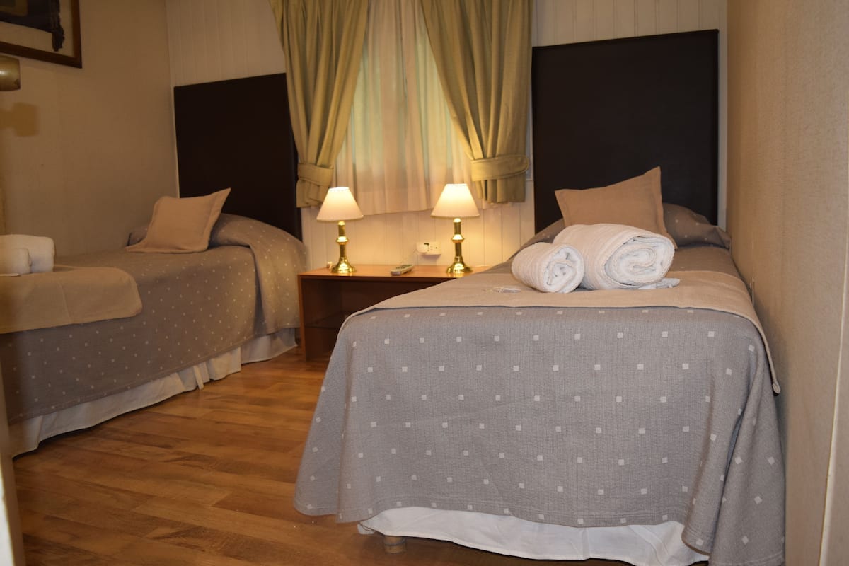Una habitación de hotel con dos camas individuales, cada una con almohada y manta. Dos lámparas de noche están sobre una mesita de madera entre las camas.