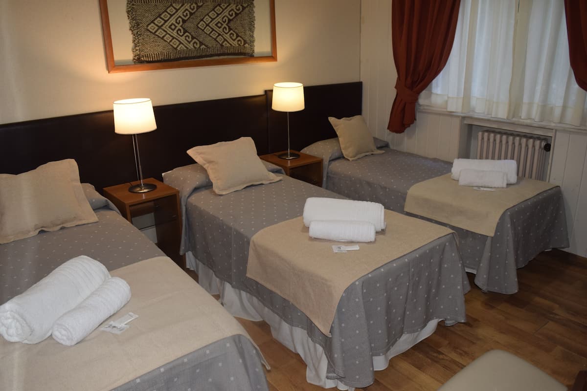 Habitación de hotel con cuatro camas individuales, cada una con almohada y toalla enrollada. Hay dos mesitas de noche con lámparas, un radiador debajo de la ventana y una obra de arte enmarcada en la pared.