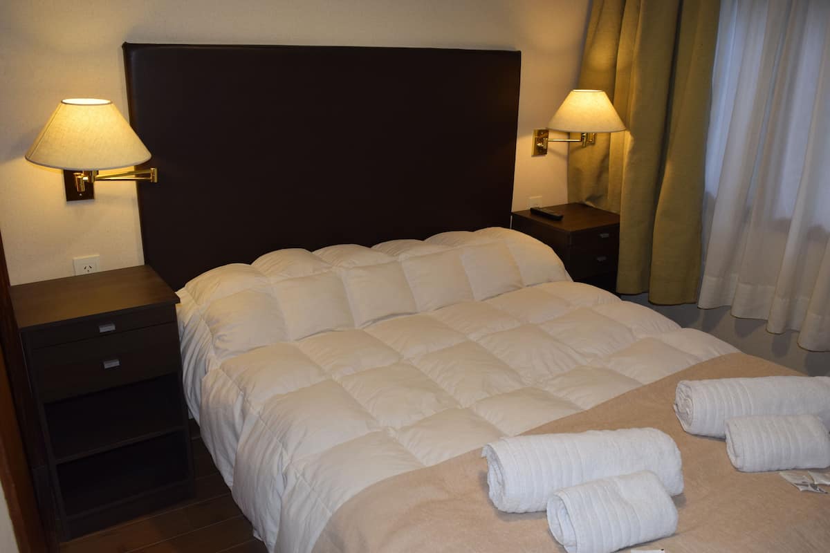 Una cama bien hecha con cabecera, dos mesitas de noche con lámparas y toallas enrolladas sobre la cama.