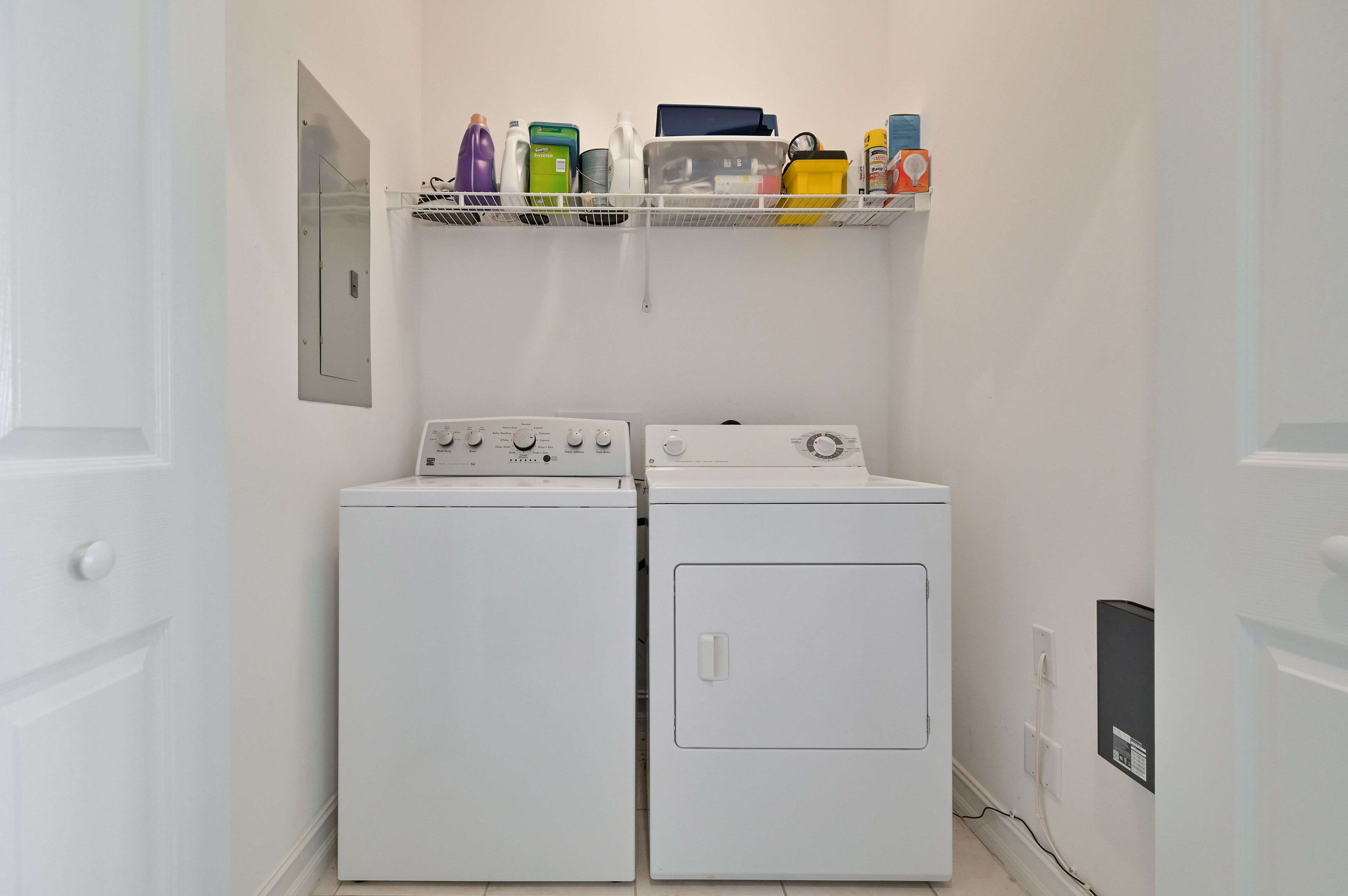 A laundry room with two white washing machines and a shelf above containing various cleaning supplies.