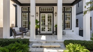 Une maison moderne avec une façade blanche, des éléments décoratifs noirs et le numéro 198 sur la porte. On y trouve un porche abrité avec deux chaises et un pot de fleurs.