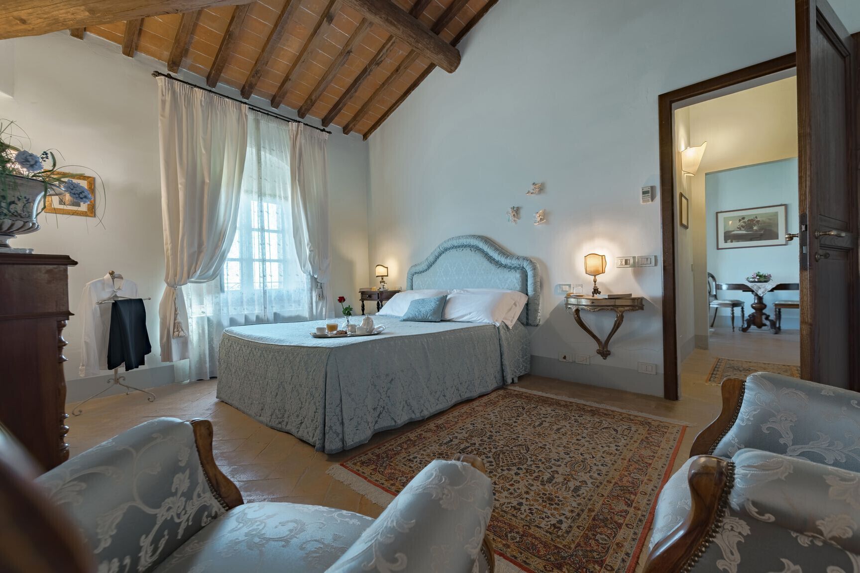 Un dormitorio con una cama grande, techo de madera, una mesita de noche con lámpara, una silla y vistas a otra habitación.