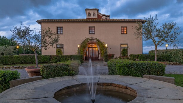A building with a central arched entrance, a fountain in the foreground, and a well-maintained garden with hedges and trees.