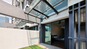 Un edificio moderno con fachada de vidrio, una pequeña zona verde y una habitación visible con cama y mesita de noche.