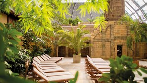 Un patio interior iluminado por el sol con hileras de sillones de madera, una planta en maceta en el centro y arquitectura de piedra.