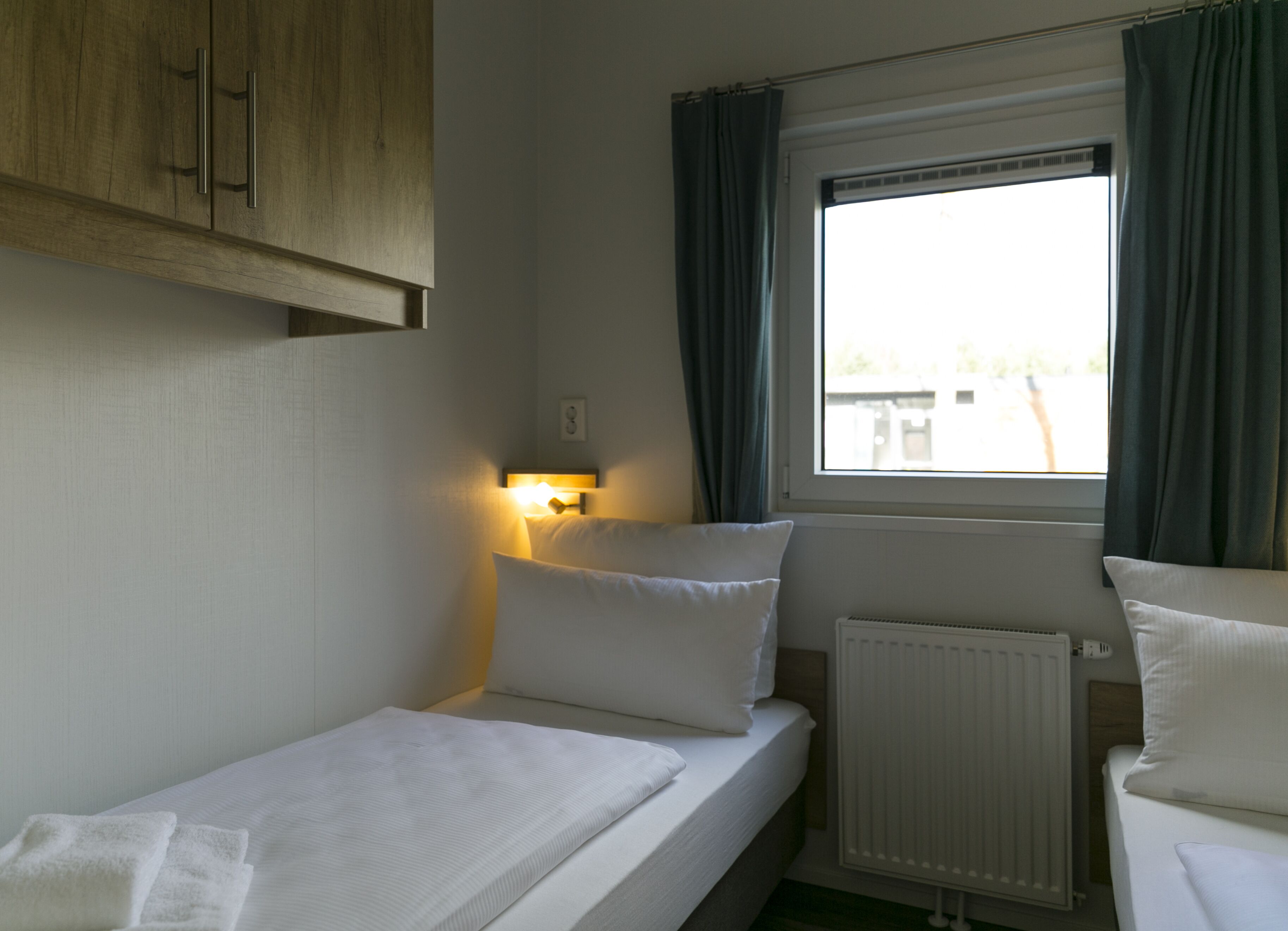 A small, single-bed room with a wooden wardrobe, a bedside table with a lamp, a window with curtains, and a radiator.