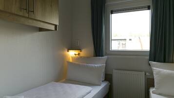 A small, single-bed room with a wooden wardrobe, a bedside table with a lamp, a window with curtains, and a radiator.