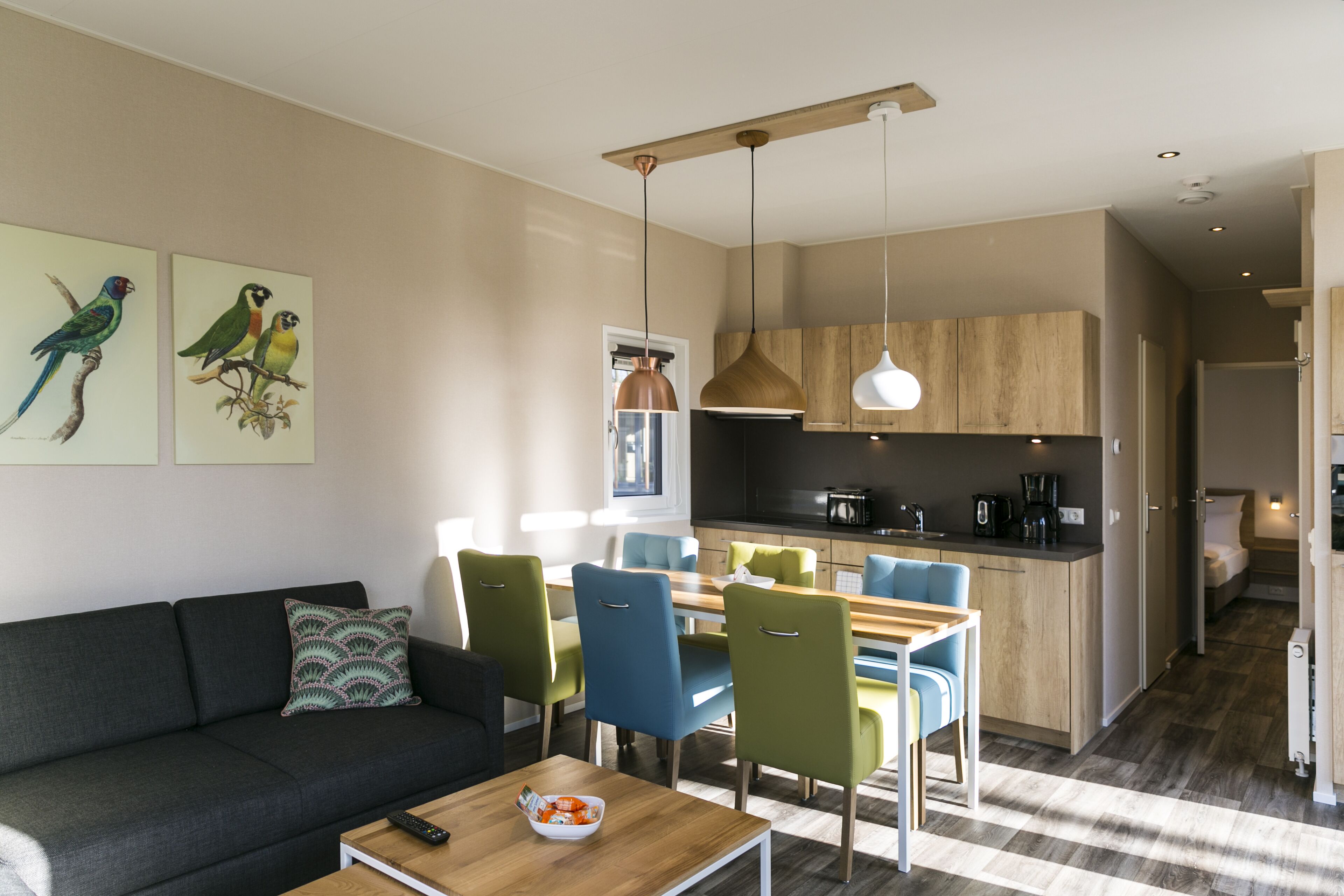 A modern living room with a sofa, a dining table with chairs, and a kitchen area with wooden cabinets and pendant lighting.