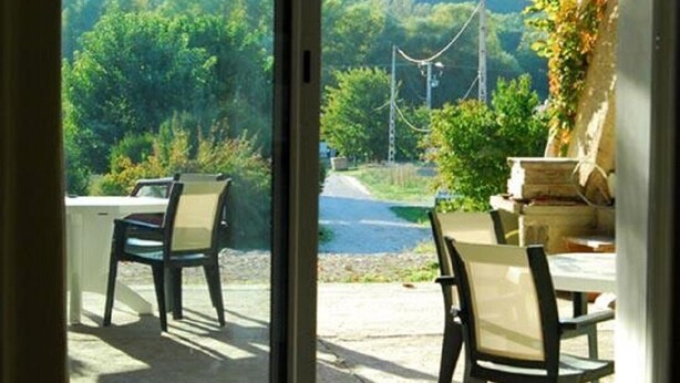 Puerta corrediza de vidrio que da a un patio exterior con dos sillas y una mesa, con vistas a un área boscosa que incluye una carretera y líneas eléctricas al fondo.