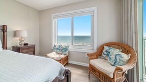 A bedroom with a large bed, a wicker chair with cushions, a nightstand with a lamp, and a window with a view of the ocean. - INCREDIABLE WEST END CAP VIEW, NICE PROPERTY, GREAT FOR FAMILIES! (Orange Beach)