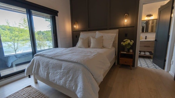 Ein Schlafzimmer mit einem großen Bett, einem Nachttisch mit Blumenvase und Seeblick durch ein Fenster.